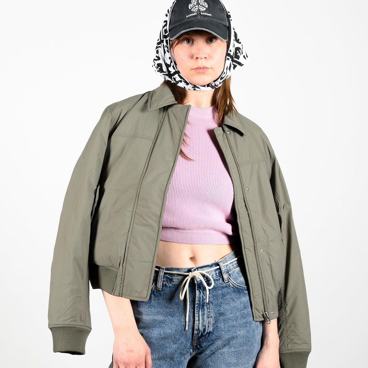 Frau im Frühlingslook mit khakifarbener Jacke, rosa Crop-Top, Jeans und gemusterter Kopfbedeckung