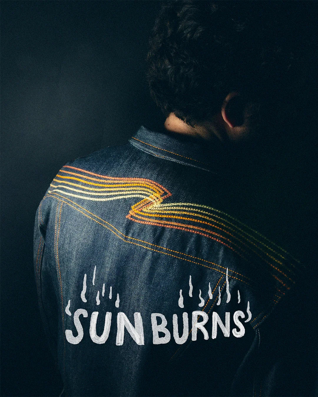 Rückansicht einer dunkelblauen Jeansjacke mit gelben Stickereien und dem Schriftzug SUNBURNS.