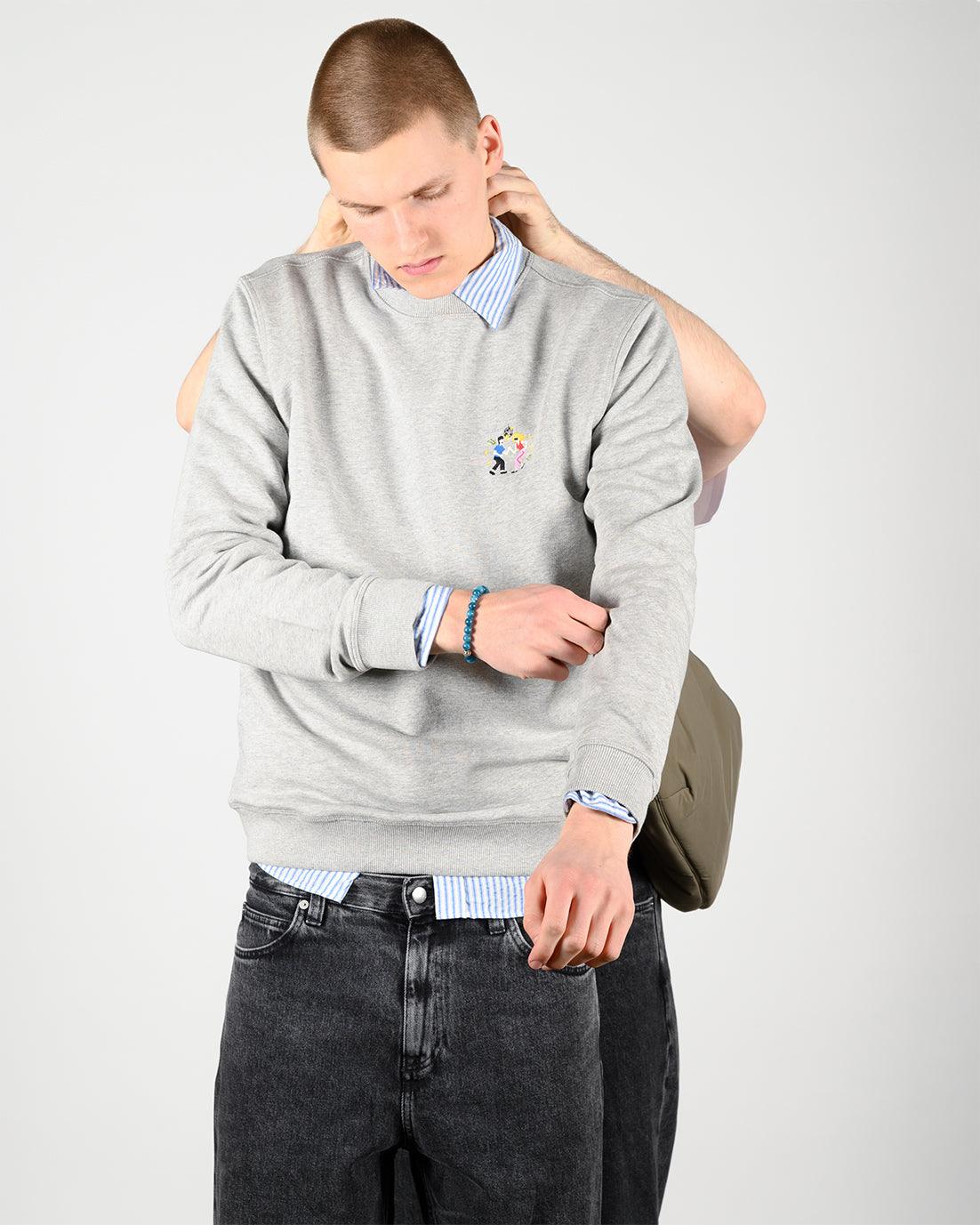 Mann in grauem Sweatshirt über blau-weiß gestreiftem Hemd und dunkler Jeans
