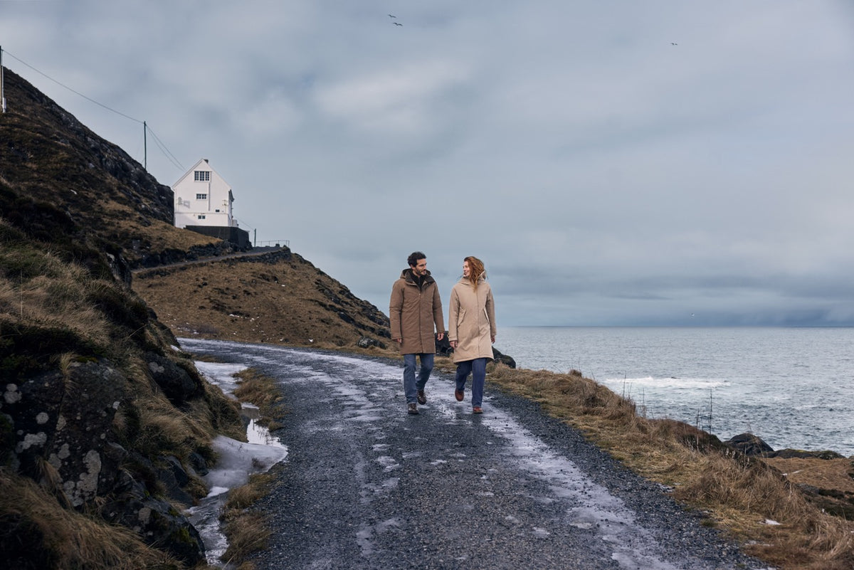 Auf dem Foto sieht man zwei Erwachsene, die beide eine Didriksons Jacke tragen, sie gehen draußen am Meer spazieren. Die Jacke haben sie im Waterkant Store in Altona gekauft und sind glücklich, weil sie spazieren gehen können ohne sich Gedanken machen zu müssen.